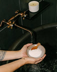 Cedar and Myrrh Sacred Bath Salts held in a ceramic bowl with a wooden scoop over a black soaking bathtub, creating a calming and intentional self-care bathing ritual.