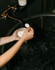 Person holding a bowl of soapy water in a dark bathroom with a candle on a shelf.