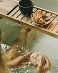 A woman in a bathtub gently kneads a bag of bath flakes under water. A ceramic cup and a burning incense stick rest on the top of the tub, creating a serene, ritual-like atmosphere.