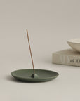 burning incense on green ceramic incense holder plate, with a ceramic bowl and book placed alongside