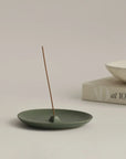 burning incense on green ceramic incense holder plate, with a ceramic bowl and book placed alongside