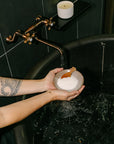 Person holding a bowl of bath salts over a bathtub with a candle on a shelf.