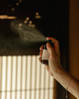 A hand spraying Hinoki Room Spray into the air, releasing a fine aromatic mist in a calm Japanese-inspired room with soft natural light.