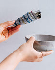hands holding white sage smudge stick wrapped in colorful string and a ceramic bowl for smudging
