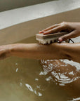 A woman in a bathtub holds a Palo Santo exfoliating bar soap and gently massages it on her arm, creating a soothing self-care moment.