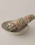 organic white sage smudge stick with red and green string resting in a ceramic bowl