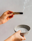 Hand holding a smoldering Palo Santo wood stick with rising smoke, over a ceramic bowl of sand used for extinguishing