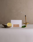 Palo Santo incense stick burning in brass holder with lemon, mint, and wood beside small box