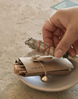 Hand holding white sage smudge bundle above Palo Santo wood, incense sticks, and brass incense holder on a ceramic bowl