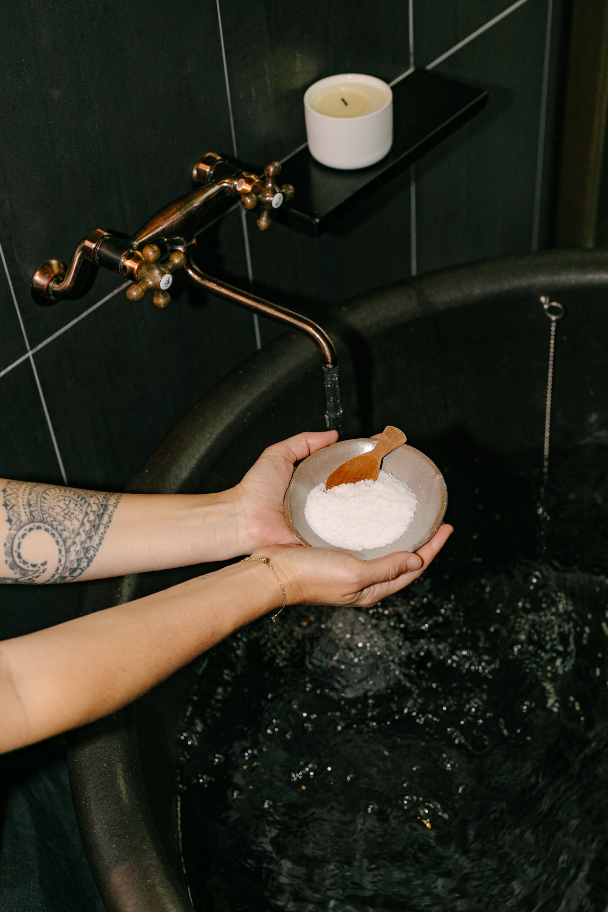 Person holding a bowl of bath salts over a bathtub with a candle on a shelf.