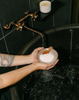 Person holding a bowl of bath salts over a bathtub with a candle on a shelf.