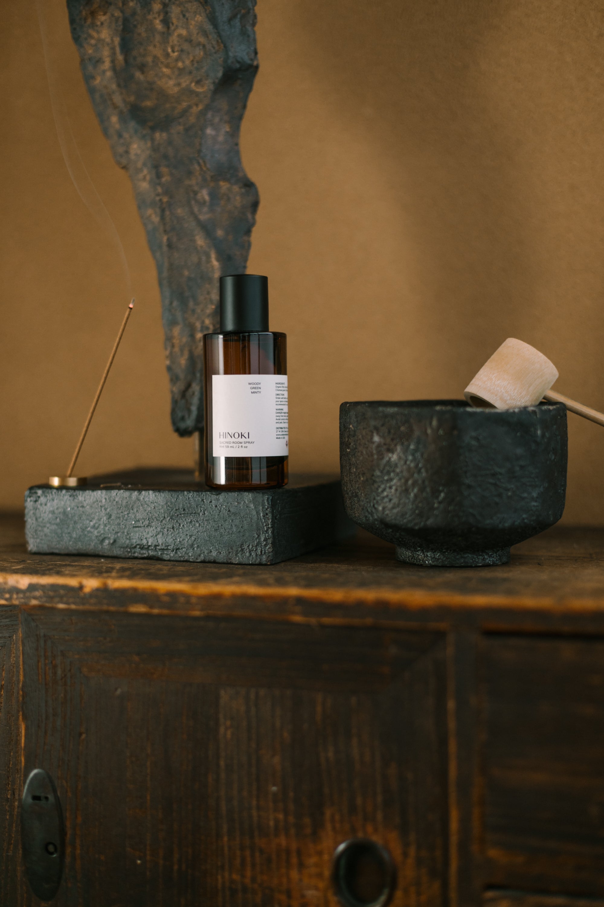 Hinoki Room Spray bottle, brass incense holder, and black ceramic bowl on a rustic wooden surface, creating a calm, minimalist ritual scene.