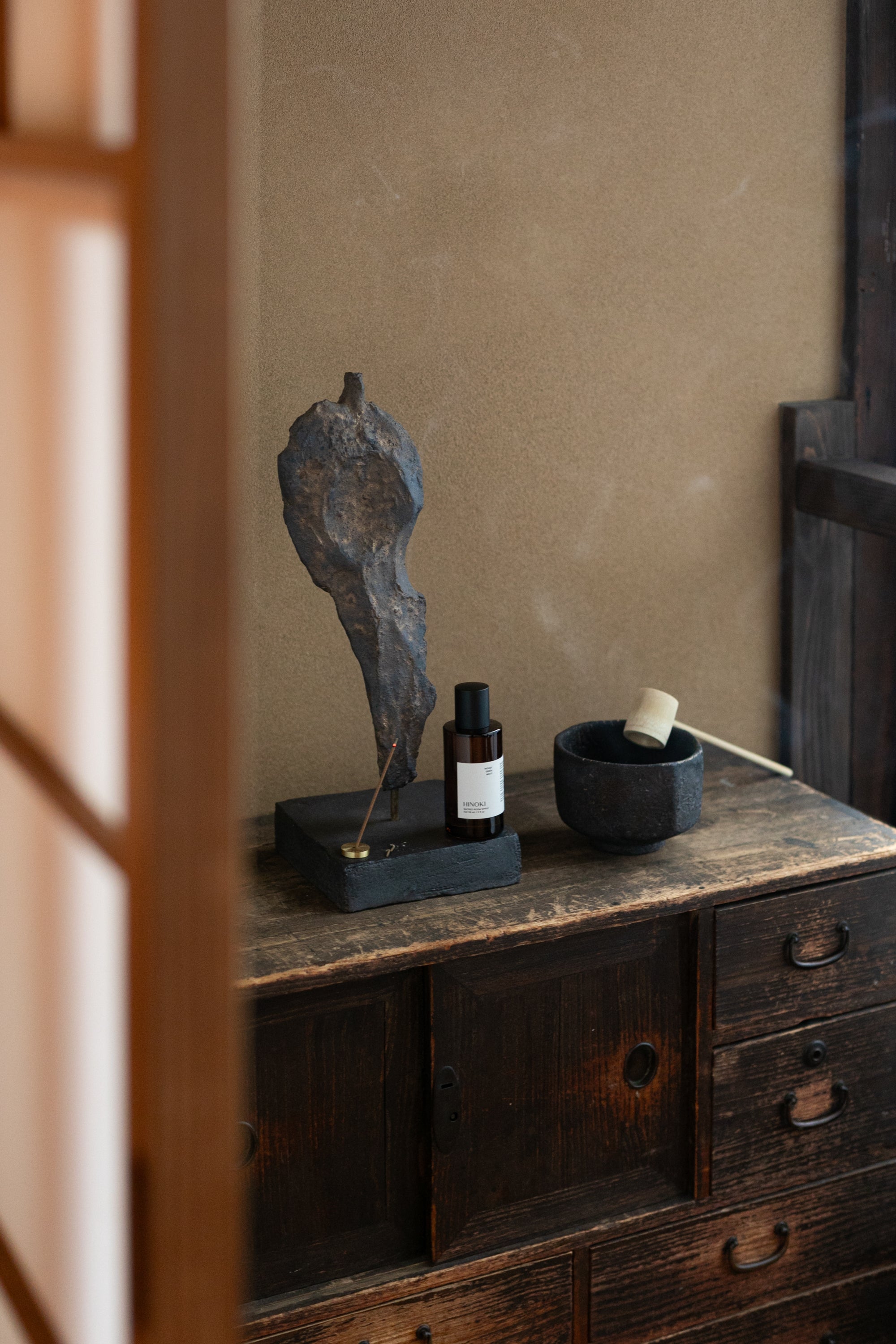 Hinoki Room Spray bottle beside a brass incense holder and black bowl on a rustic Japanese wooden cabinet, evoking calm and mindful living.