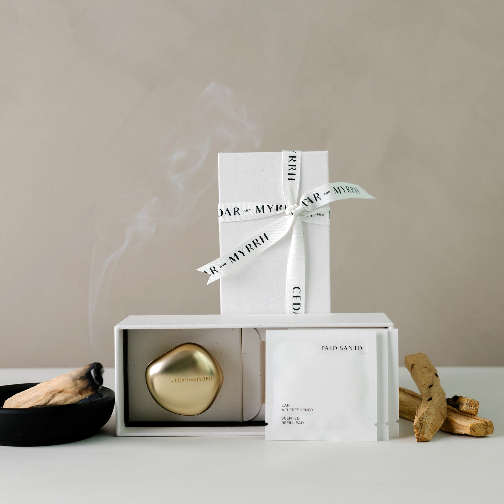 Decorative items including a gold car air refreshener, white boxes with ribbons, and Palo Santo sticks on a neutral background.