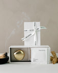 Decorative items including a gold car air refreshener, white boxes with ribbons, and Palo Santo sticks on a neutral background.